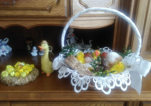 Wiklinowy koszyk wielkanocny stoi na kredensie. obok stoi gliniana kaczka w kapeluszu i żółte kurczaczki na sianku.