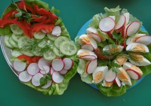 Dwa talerze z wielkanocnym śniadaniem. Na prawym talerzu ułożone są ćwiartki jajek, na lewym talerzy ułożone są warzywa: zielone ogórki, rzodkiewka i pomidor ozdobione koprem i natką pietruszki