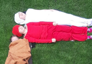 Troje dzieci leży na trawie, tworzą flagę. Dwoje dzieci leży poziomo. Górne dziecko ubane jest na biało, dolne dziecko ubranee jest na czerwono. Trzecie dziecko leży pionowo, ubranie ma w kolorze beżowym.