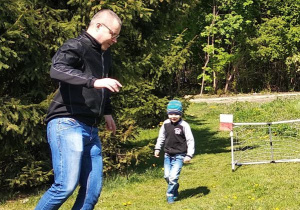 Tato i syn grają na murawie w piłkę nożną. W prawym górnym rogu bramki powiewa flaga biało - czerwona