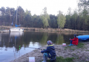 Dwoje dzieci ubrane w kurtki i czapki siedzi na stołeczkach nad zalewem, w dłonich trzymaja wędki częściowo zanurzone w wodzie i łowią ryby.