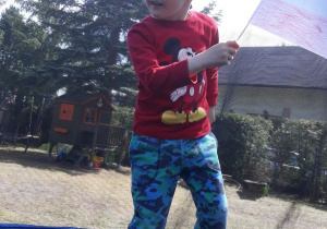 Chłopiec skacze na trampolinie, w ręęce trzyma choragiewkę biało - czerwoną.