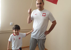 Tato i syn ubrani w koszulki białe z herbem Polski stoją w pustym pokoju. Tato trzyma w prawej ręce flagę polski. Syn dotyka lewą stopą kolorową pilkę.noga piłkę