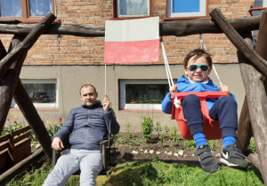 Tato i syn przed domem huśtają się na huśtawkach. Tato w lewej dłoni trzyma flagę Polski