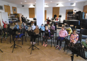 Dzieci siedzą na krzesełkach gdzie odbywają się próby orkiestry.