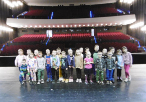 Dzieci stoją na wielkie scenie teatru, w tle widać sale widowni.