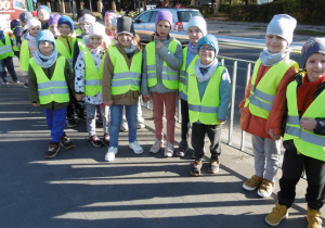 Dzieci z grupy 5 stoją na przystanku tramwajowym, są ubrane w kamizelki odblaskowe.