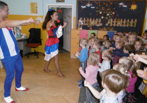 Dzieci stoją w sali na dywanie i naśladują ruchy tancerzy.