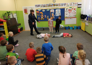 Dzieci prezentują scenkę, dwoje leży na dywanie udając nieprzytomnych wyznaczony chłopie i dziewczynka stoją nad nimi i pokazują jak należy zachować sie w takiej sytuacji.