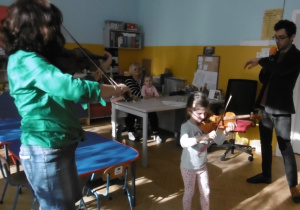 Dziewczynka trzyma skrzypce i próbuje grać , obok ze swoimi skrzypcami stoi pani Bożenka i prowadzący.