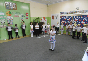 Przedszkolaki stoją na dywanie, pośrodku dziewczynka mówi przez mikrofon.