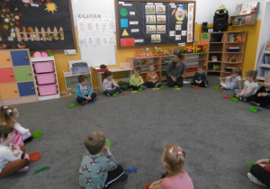 dzieci siedzą w kole na dywanie, przed nim są pojemniczki i kolorowe figury geometryczne..