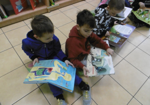 Dzieci siedzą na podłodze i prezentują ilustracje w książkach.