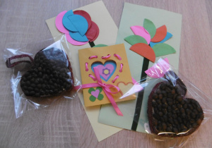Na stoliku leżą prezenty dla dziadków: laurka z kwiatkiem, kolorowe serduszko i zapakowane ciasteczko w kształcie serca.
