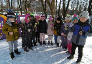 Dzieci stoją w ogrodzie przedszkolnym i trzymają w rękach zawieszki z ziarnem dla ptaków.
