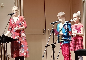 Na scenie stoi nauczycielka ze szkołymuzycznej z dwójką dzieci.