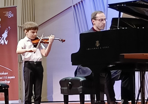 Na fortepianie gra nauczyciel, obok stoi chłopiec i gra na skrzypcach.