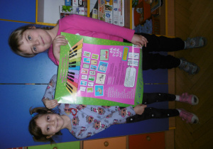 Dwie dziewczynki w sali przedszkolnej trzymają plakat ze szkoły muzycznej.