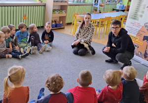 Dzieci z grupy 1 siedzą w kole na dywanie. Prowadzące osoby prezentują prelekcję.