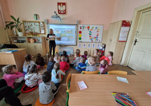 Dzieci siedzą na podłodze przed tablicą. Nauczycielka pokazuje książkę związaną z tematyką zajęć.