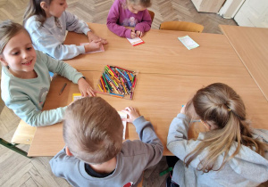Przedszkolaki siedzą przy stolikach i wykonują prace plastyczne przy użyciu kredek.