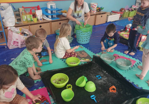 Dzieci siedzą na rozłożonych ceratach na których leża narzędzia i stoją naczynia z ciastem plastycznym