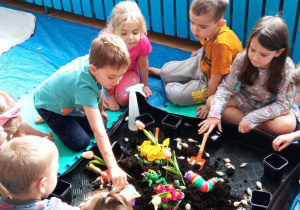 Dzieci siedzą na rozłożonych ceratach i eksperymentują z ziemią i roślinami. obok stoja małe czarne ddoniczki