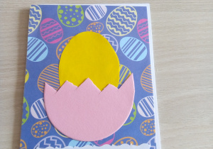 Pocztówka wielkanocna. Żółte jajko w różowej połówce skorupki na niebieskim tle z kolorowymi pisankami. Na dole pocztówki napis wesołych świąt