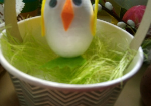 Kurczak z białego jajka styropianowego siedzi na zielonym sianku w koszyczku z pałączkiem