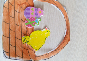 Papierowy ,żółty kurczak stoi w papierowym koszyku. Przed kurczakiem dwie kolorowe pisanki