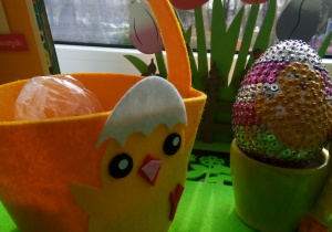 Pomarańczowy koszyk z materiału, ozdobiony żółtym kurczakiem ze skorupką na głowie. Po prawej stronie stoi doniczka z jajkiem ozdobionym cekinami