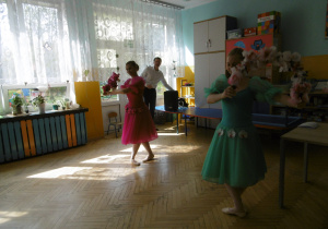 Dwie baletnice. Po lewej Po lewej stronie stoi na puentach baletnica w różowej sukni. Po prawej stronie stoi na puentach baletnica w turkusowej sukni. Baletnice trzymają w dłoniach wianki z kolorowych kwiatów.