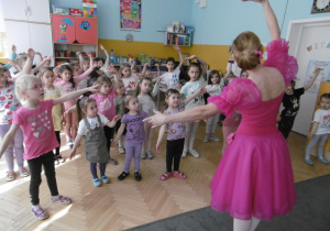 Dziewczynki tańczą układ baletowy pokazywany przez baletnicę w różowej sukience. Baletnica stoi twarza do dzieci.