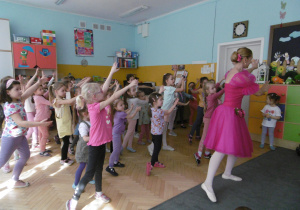 Dziewczynki tańczą układ baletowy pokazywany przez baletnicę w różowej sukience. Baletnica stoi przed dziećmi, odwrócona plecami