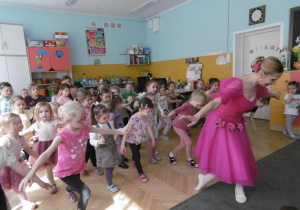 Dziewczynki ćwiczą ukłon pokazywany przez baletnicę w różowej sukience. Baletnica stoi przed dziećmi, odwrócona plecami