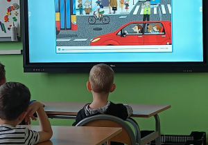 Plansza ulicy na tablicy multimedialnej. Chłopcy siedza w ławkach i patrzą na ekran tablicy.