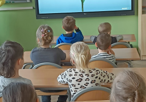 Dzieci siedza w ławkach szkolnych i patrzą na obraz kwiatka na tablicy multimedialnejSzkoła11