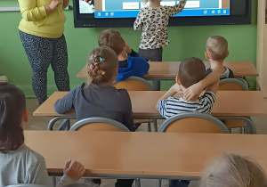 Dzieci siedza w ławkach szkolnych. Nauczycielka stoi obok tablicy. Dziewczynka zaznacza na tablicy części kwiatka.