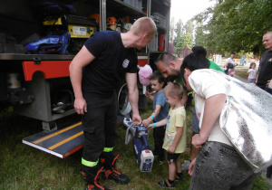 Dzieci oglądaja wyposażenie wozu strażackiego. Pan strażak opowiadda do czego dany przyrzą służy