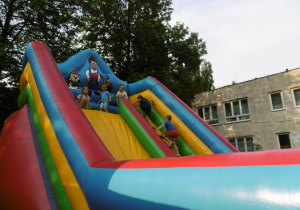 Troje dzieci stoi na szczycie dmuchańca - zjeżdżalni. Przygotowują sie do zjazdu. Po prawej stronie dziewczynka i chłopiec wchodzą na górę zjeżdzalni