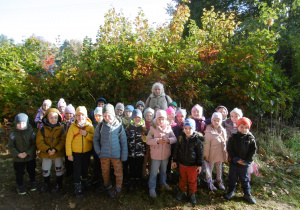 Grupa IV pozuje do zdjęcia razem z wychowawczynią, na tle kolorowych jesiennych krzewów.