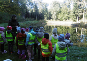 Dzieci wraz z leśniczym stoją nad brzegiem leśnego jeziora, słuchając opowieści o bobrach i ich zwyczajach.