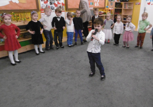 Chłopiec w białej koszuli trzyma w dłoniach mikrofon i śpiewa piosenkę.