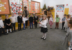 Dzieci stoją w półkolu. Na środku stoi dziewczynka, w dłoni trzyma mikrofon i recytuje wiersz.