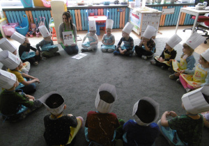 Dzieci-cukiernicy siedzą na dywanie w kole. Pani dietetyczka prezentuje historyjkę obrazkową o zdrowym żywieniu.