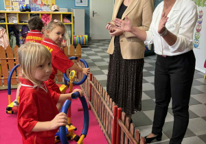 Troje dzieci ćwiczy na przyrządach w mini siłowni. Przed dziećmi stoi pani wiceprezydent miasta Łodzi i pani dyrektor przedszkola 156.